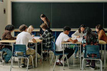 Une salle de classe. Il y a deux groupes d'élèves. Leurs bureaux sont placés ensemble pour travailler en équipe. Une enseignante marche entre les deux groupes d'élèves.
