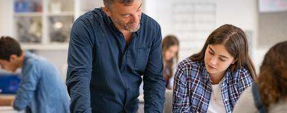 Dans une salle de classe, les élèves écrivent à leur pupitre. Un enseignant et une élève regardent ensemble un devoir.