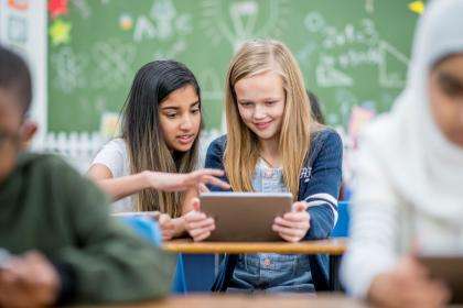 Dans une salle de classe, les élèves sont assis à leur pupitre. Deux élèves font une activité d'apprentissage à l'aide d'une tablette.