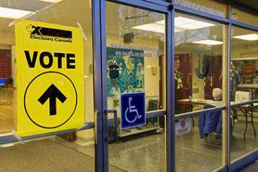 Fenêtres et porte d'entrée d'un bureau de vote. Sur une vitrine, une affiche jaune lit: "vote". À l'intérieur du bâtiment il y a une table d'enregistrement. Une affiche d’un fauteuil roulant est accrochée à la porte.