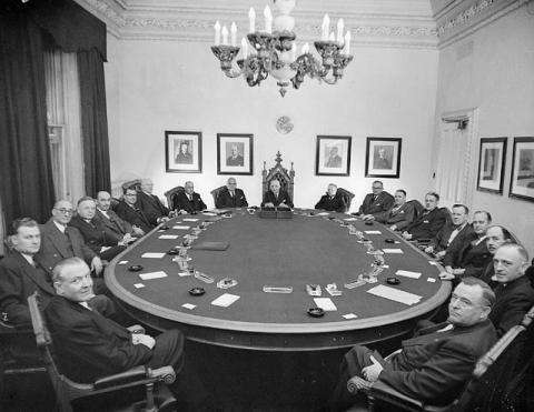 Photographie en noir et blanc d’une réunion de cabinet. Un groupe de parlementaires est assis autour d’une grande table ovale. 