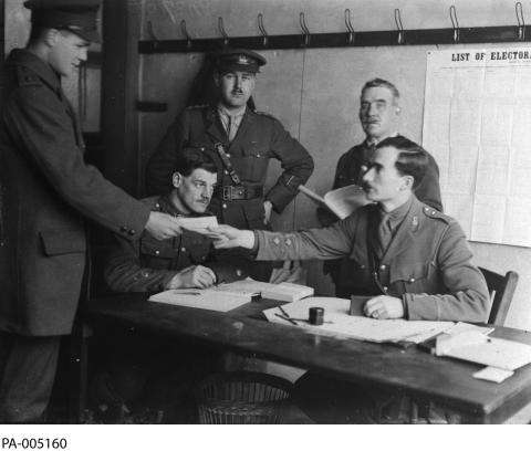 Photographie en noir et blanc d’un groupe d’hommes en uniforme militaire, rassemblés autour d’une table. Un homme tend un bulletin de vote à un autre. Au mur, une affiche indique en anglais: « List of Electors » La traduction : « Liste électorale ».