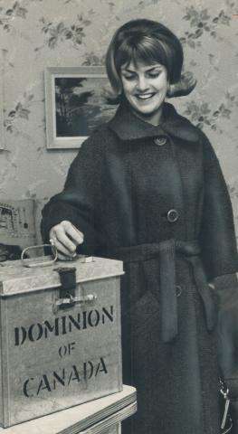 Photographie en noir et blanc d’une jeune femme souriante plaçant son bulletin de vote dans une urne en métal.