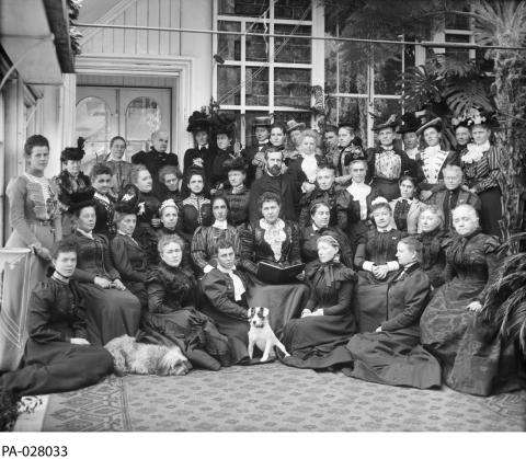 CE13-Women_FR Photographie en noir et blanc de plusieurs rangs de femmes. Deux chiens sont assis à l’avant.