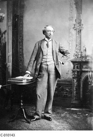 CE14-Women_FR Photographie en noir et blanc de Sir John A. MacDonald debout devant un foyer, une main sur la hanche, l’autre touchant un livre ouvert sur une petite table à côté de lui.