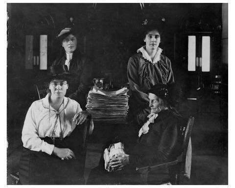 CE15-Women_FR Photographie en noir et blanc de quatre femmes assises autour d’une table sur laquelle est posée une grosse pile de papier.