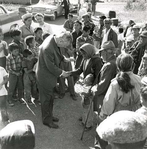 CE19-Women_FR Photographie en noir et blanc du premier ministre John Diefenbaker serrant la main d’une femme autochtone, parmi un petit groupe de personnes de tous âges. La femme entoure un jeune enfant du bras.
