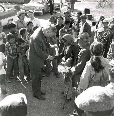 Photographie en noir et blanc du premier ministre John Diefenbaker serrant la main d’une femme autochtone, parmi un petit groupe de personnes de tous âges. La femme entoure un jeune enfant du bras.