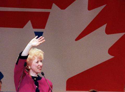 Photographie de Kim Campbell debout derrière un microphone, faisant des signes de la main en souriant.