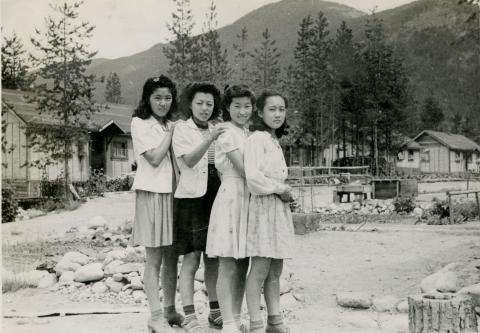 Carte contextuelle : les Canadiens d’origine japonaise Photographie en noir et blanc de quatre jeunes femmes japonaises se tenant par les épaules devant un camp d’internement, avec des montagnes en arrière-plan.