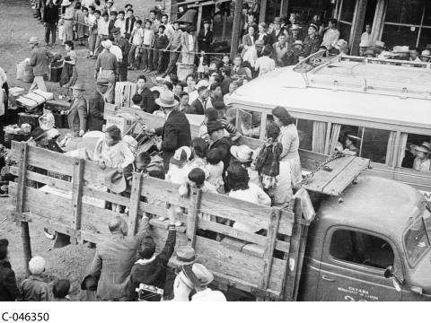 Photographie en noir et blanc d’un grand groupe de personnes de tous âges embarquant à l’arrière d’un camion avec leurs bagages. Une longue file attend aussi derrière une barrière de corde.