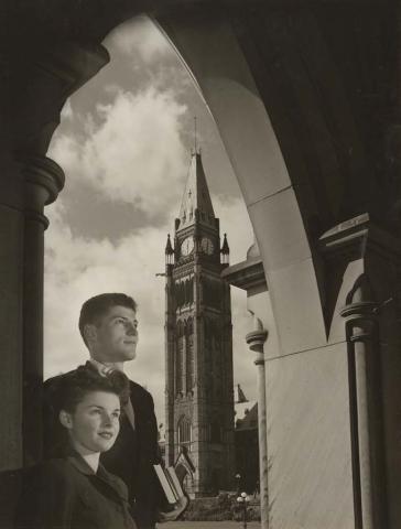 Photo en noir et blanc de deux jeunes personnes en tenue des années 1960. Elles se tiennent debout devant le Parlement, avec la tour de la Paix en arrière-plan.