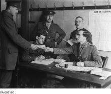 CE04-Youth_FR Photographie en noir et blanc d’un groupe d’hommes en uniforme militaire, rassemblés autour d’une table. Un homme tend un bulletin de vote à un autre. Au mur, une affiche indique en anglais: « List of Electors » La traduction : « Liste électorale ».