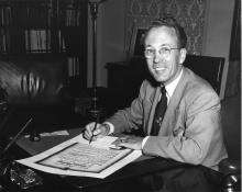 CE05-Youth_FR Photographie en noir et blanc de Tommy Douglas assis à son bureau, signant un document officiel et souriant à la caméra.
