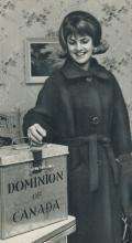 CE11-Women Photographie en noir et blanc d’une jeune femme souriante plaçant son bulletin de vote dans une urne en métal.