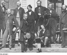 Photographie en noir et blanc d’un groupe d’hommes, incluant Sir John A. MacDonald, rassemblés sur des marches et tenant leur chapeau haut de forme.