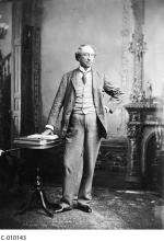 Photographie en noir et blanc de Sir John A. MacDonald debout devant un foyer, une main sur la hanche, l’autre touchant un livre ouvert sur une petite table à côté de lui.