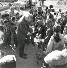 CE19-Women_FR Photographie en noir et blanc du premier ministre John Diefenbaker serrant la main d’une femme autochtone, parmi un petit groupe de personnes de tous âges. La femme entoure un jeune enfant du bras.