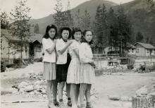 Photographie en noir et blanc de quatre jeunes femmes japonaises se tenant par les épaules devant un camp d’internement, avec des montagnes en arrière-plan.