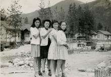 Carte contextuelle - Canadiens d'origine japonais Photographie en noir et blanc de quatre jeunes femmes japonaises se tenant par les épaules devant un camp d’internement, avec des montagnes en arrière-plan.