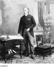 CE34-Japanese_FR Photographie en noir et blanc de Wilfrid Laurier debout à côté d’une table, la main posée sur un grand livre ouvert.
