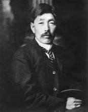 Plan taille en noir et blanc d’un homme japonais moustachu tenant un chapeau dans ses mains.