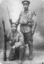 Photographie en noir et blanc de deux hommes japonais en uniforme militaire tenant deux fusils à leur côté. Un homme est debout et l’autre s’appuie sur un genou devant lui.